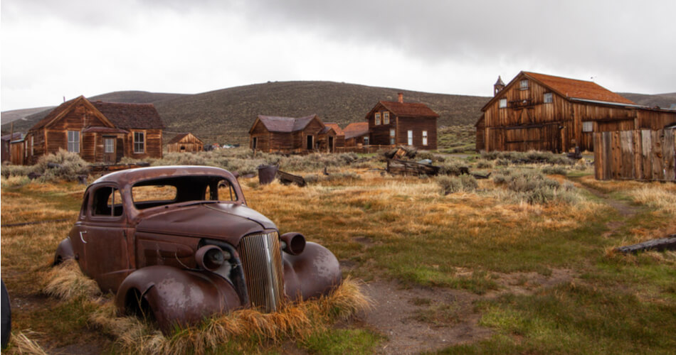 Old rusted car sitting in a field in front of dilapidated buildings